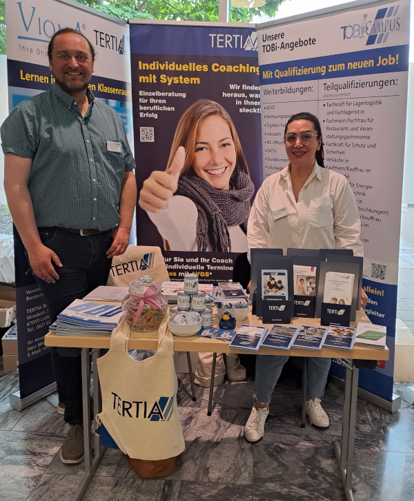 Zwei Mitarbeiter:innen stehen hinter einen Messestand, der mit Werbeartikeln bestückt ist.